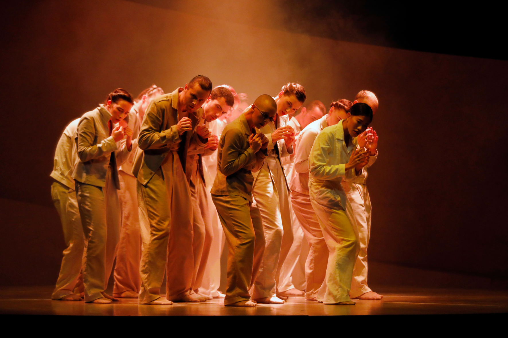 Een groep dansers met handen als vuisten voor de borst, ietwat gebogen en ingetogen dansend in een oranje licht.