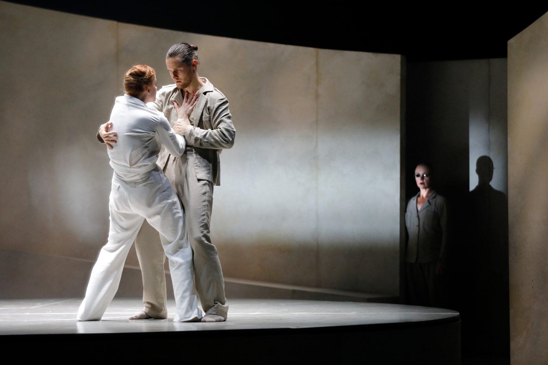 Two dansers in duet on the stage. We see the back of a female, who has her hand against the chest of her partner. He has his hand around her wrist and the other arm on her back. A mysterious figure is watching in the background.