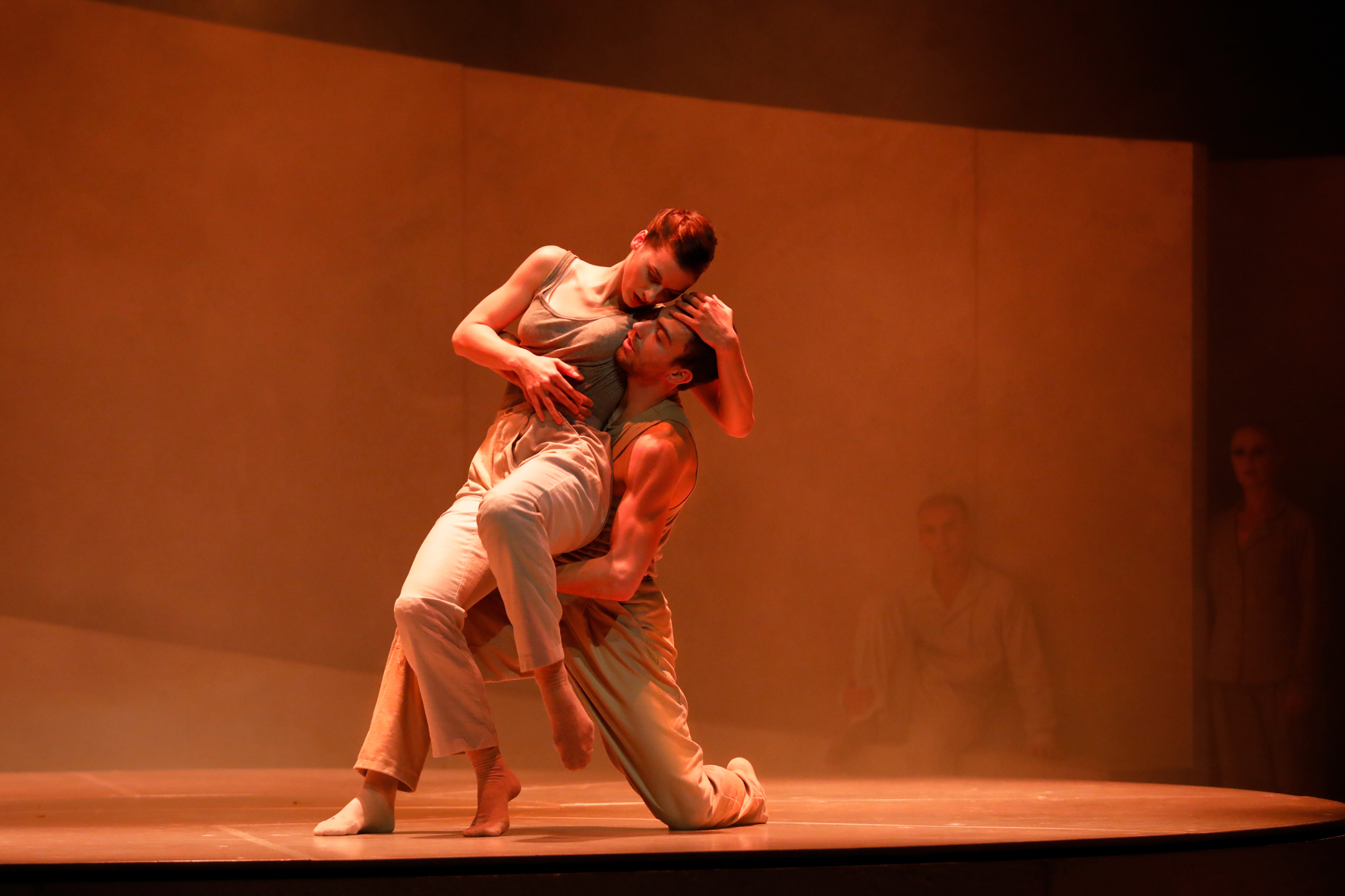 A male and female dancer in a light suit on a stage in orange lights. He is on one knee and she leans on him and has his head in her hands.