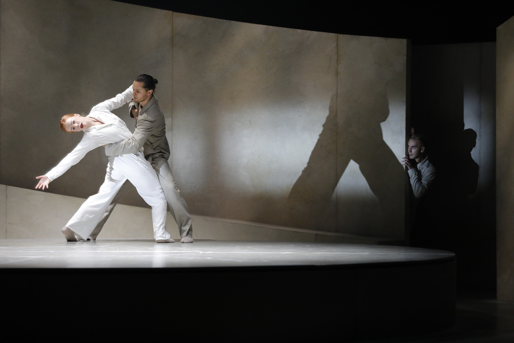 Two dancers on stage. The male dancer has his arm around her back as she leans backwards and had her arm stretched back. At the background someone is watching from the shadow.