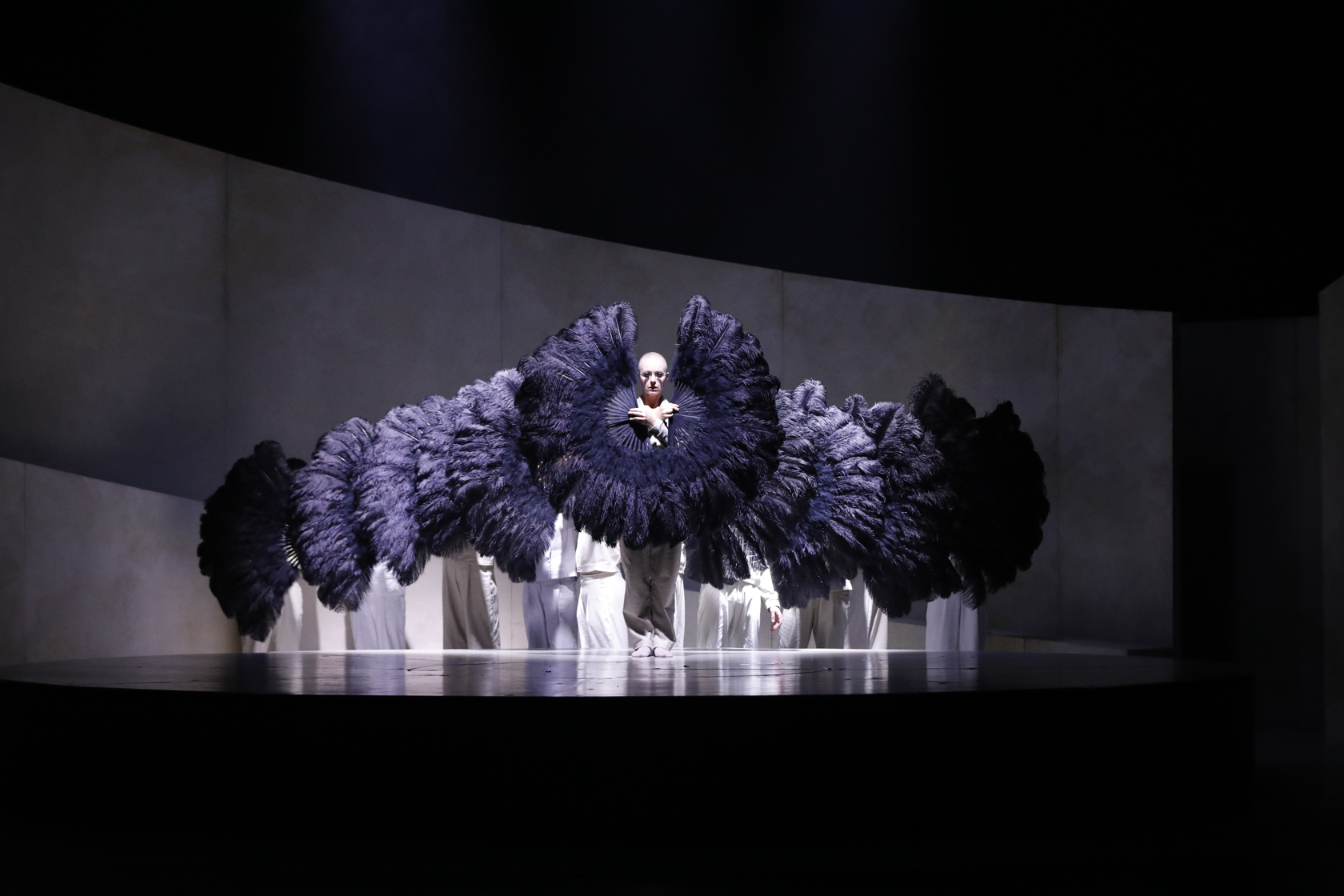Dancers in a triangle shape on stage, but we only see the face of the first. All dancers have two big fans with black feathers in their hands. 
