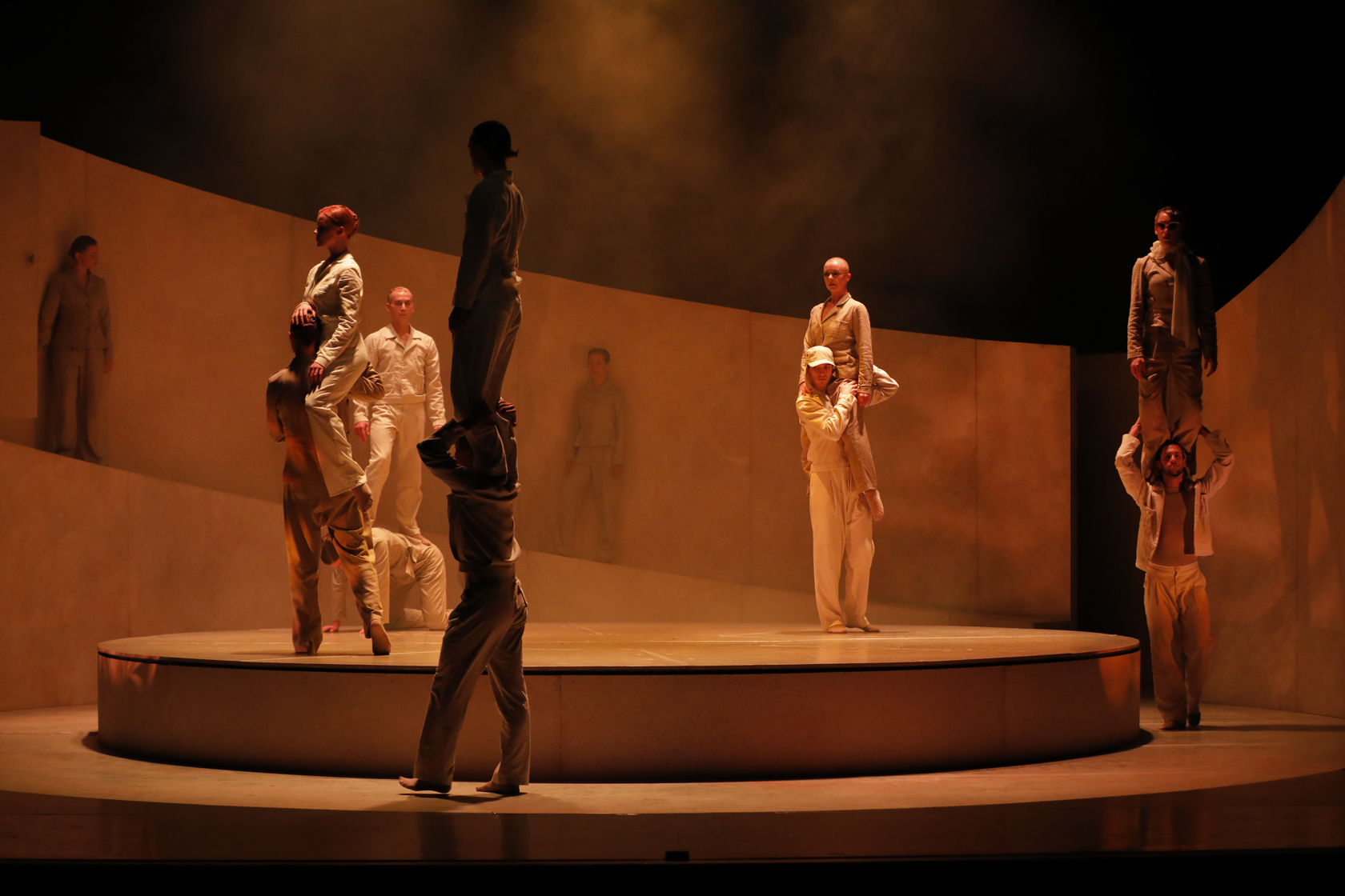 Dansers op en rondom een rond podium in oranje licht met rook. Zij lopen rond en dragen een andere danser op de schouders.