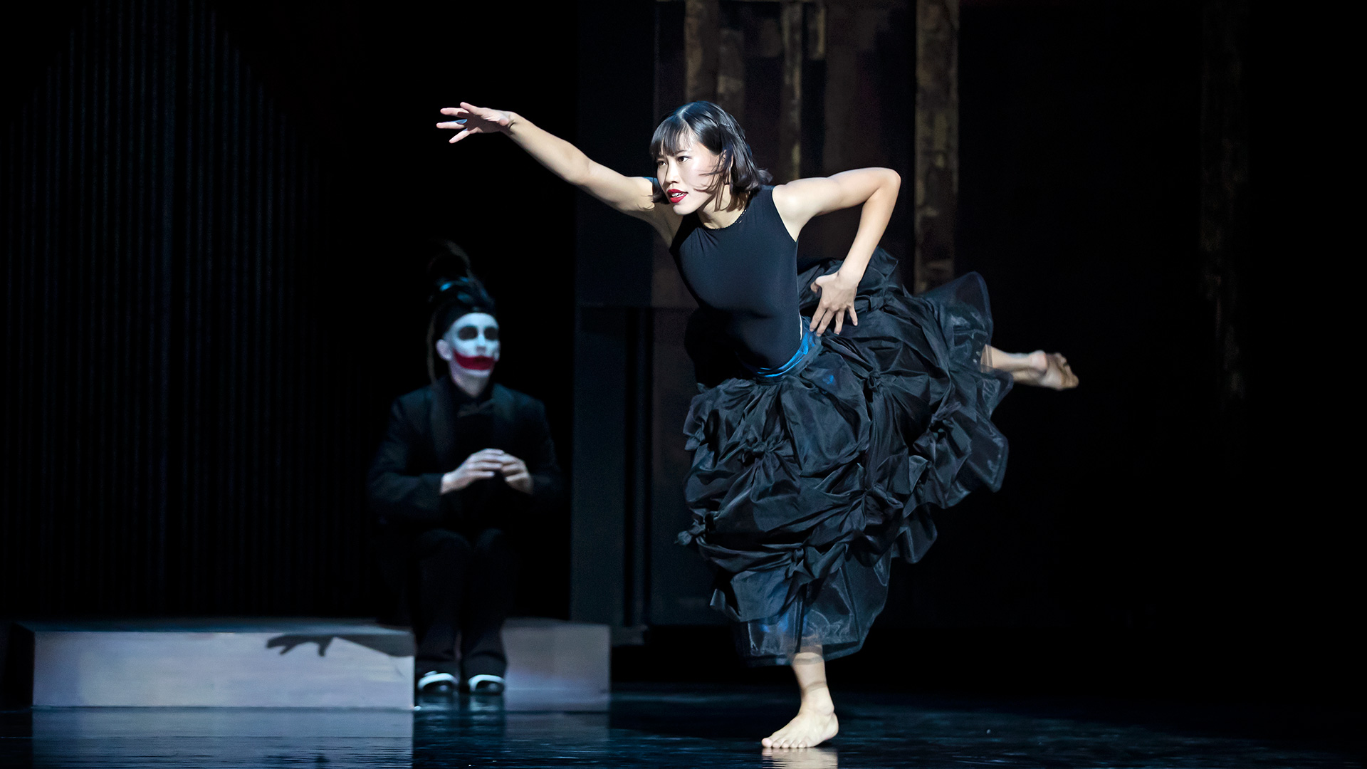 Danseres Sana met uitgestrekte arm en been in zwarte jurk in twenties stijl, met op de achtergrond een geschminkte Mischa