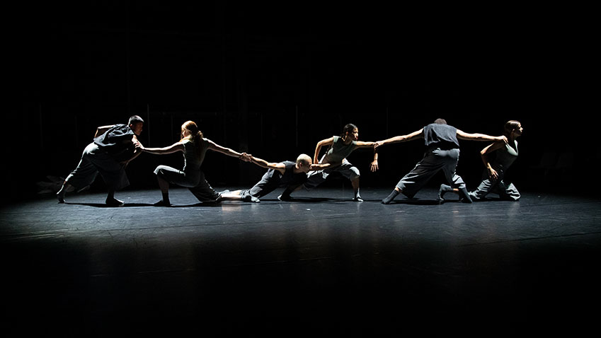 Zes dansers gekleed in het grijs, die elkaar vasthouden met de armen