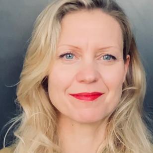 Blonde vrouw met golvend jaar, helderblauwe ogen en roodgestifte lippen met een glimlach. Ze staat tegen een grijsblauwe achtergrond.