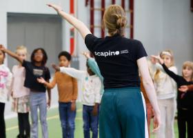 Vrouw gezien vanaf de achterkant met een zwart shirt met daarop een wit Scapino logo. Zij staat in een gymzaal met een klas vol leerlingen die hun arm omhoog hebben gestoken.