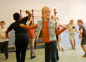 Klas met lachende kinderen in een gymgaal die enthousiast dansen met hun armen opzij