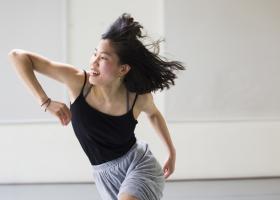 Danseres met donker halflang haar dat naar achter wappert in een lichte studio. Zij heeft een elleboog gebogen en schuin omhoog. Haar andere arm is naar achteren.