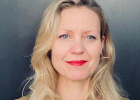 Blonde vrouw met golvend jaar, helderblauwe ogen en roodgestifte lippen met een glimlach. Ze staat tegen een grijsblauwe achtergrond.