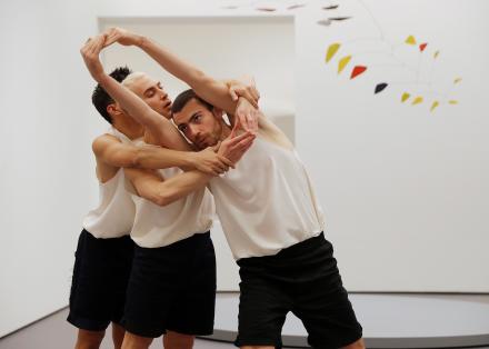 Drie dansers met zwarte broek en een wit hemd. Zij staan in een witte omgeving, waarin we rechts bovenin een fragiele mobile zien van ijzerdraad en gele, rode en zwarte driehoekjes. De dansers zijn met de armen in elkaar gevlochten.