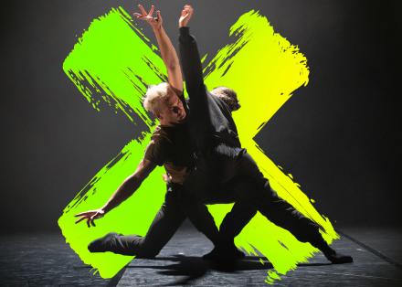 Twee dansers in het zwart met armen geopend en geknield op de vloer. Op de achtergrond een neongeelgroen kruis.