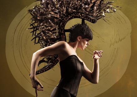 Woman in a black corset with bare arms and a large gold headdress in the shape of a horse's mane. She has raised her arms gracefully as if walking. The background is gold with a painted circle.